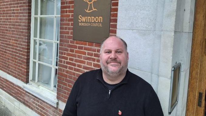 Jim Robbins stands outside the Swindon Borough Council office. He has short grey hair and stubble and wears a black half-zip jumper.