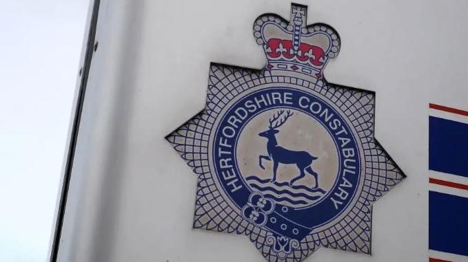 Large emblem of Hertfordshire Constabulary set on a building. It is blue and white and features the blue hart - a male deer -  of Hertfordshire in the centre of the emblem. Above the hart is a red crown. The symbol is set against an off white background and there is a section of cloudy grey sky to the left of the police logo. 