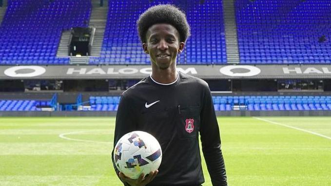 A man wearing a black referee kit while holding a football. He is standing on the pitch at Portman Road stadium. He is looking into the camera. 