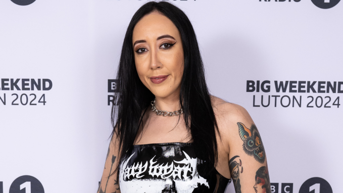 Alyx Holcombe wearing a black top with white writing on it. She has tattoos on both arms, black hair and black jeans.