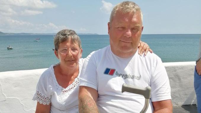 A lady and man sit together in front of a white wall with the blue sea behind. The lady has short grey hair, glasses a white frilly top and has her left arm around the man. The man has short white hair and is wearing a white T-shirt with Berghaus written on it and carrying a walking aid. They are looking at the camera and smiling. 