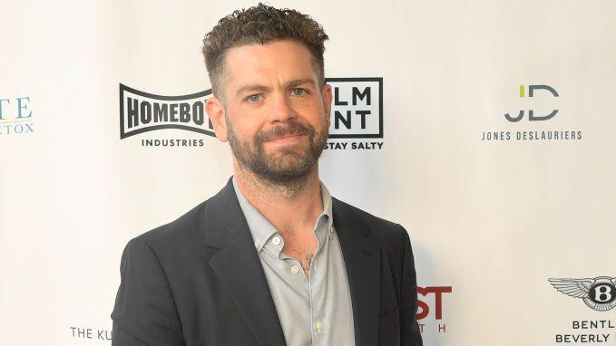 Jack Osbourne, who has dark hair cropped short and a beard, standing in front of a hoarding bearing various company logos. He is wearing a grey suit jacket and pale grey shirt and smiling.