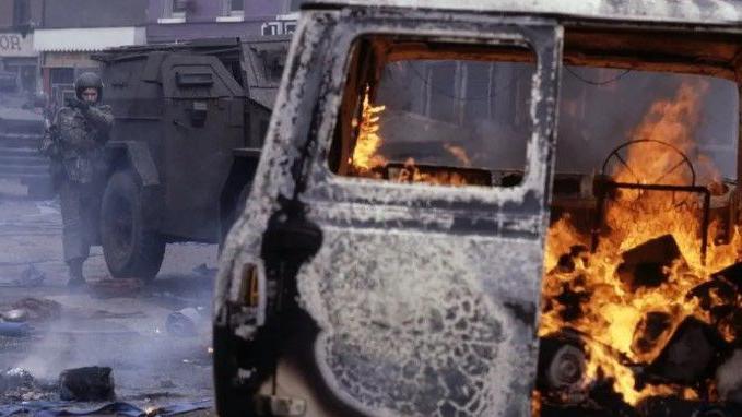 An armed British Army soldier walks toward a burning van beside a large armoured vehicle