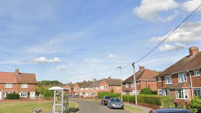 A residential street with houses, parked cars, a bus stop and grass verges.