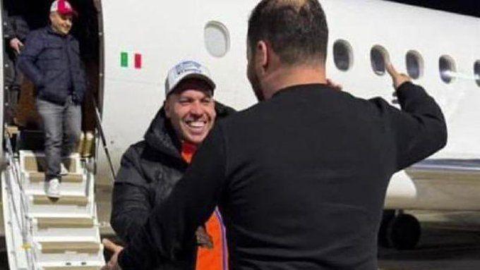 A man embraces another man after getting off a small passenger jet with an Italian flag on the side. The man facing the camera is smiling. He's wearing a black jacket and an orange T-shirt.