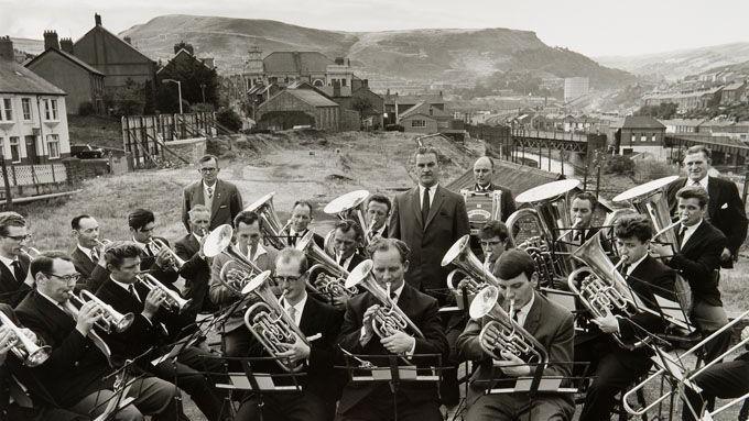 Mid-Rhondda Workman's Band in Penygraig in 1964: a black and white photo showing a large group of men in suits playing brass instruments, with the valley, hills and housing in the background
