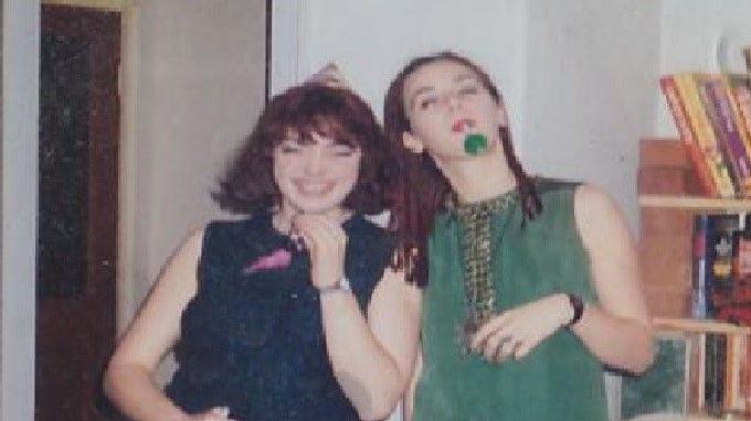 Two women face the camera. The one on the right is wearing a green dress and blowing a party popper. The one on the left is in a navy blue vest top and is laughing.