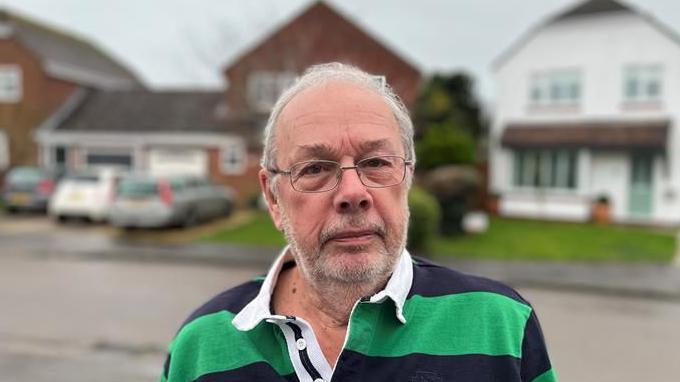 Ray Palmer looking solemnly at the camera standing on the pavement where the flooding was on the road behind him. There are three detached houses behind him with cars on the driveway and Ray is wearing a green and black stripey top and has glassed on 