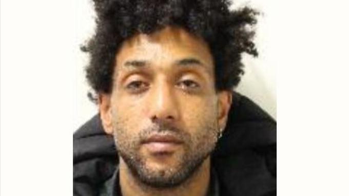 Low-res image of Dante Forrest in a police custody mugshot. He wears a black puffa jacket, has curly hair and an earring.