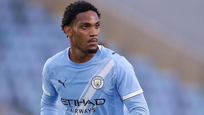 Christian McFarlane wearing a light blue Manchester City shirt. There are blue seats in a stadium behind him. 