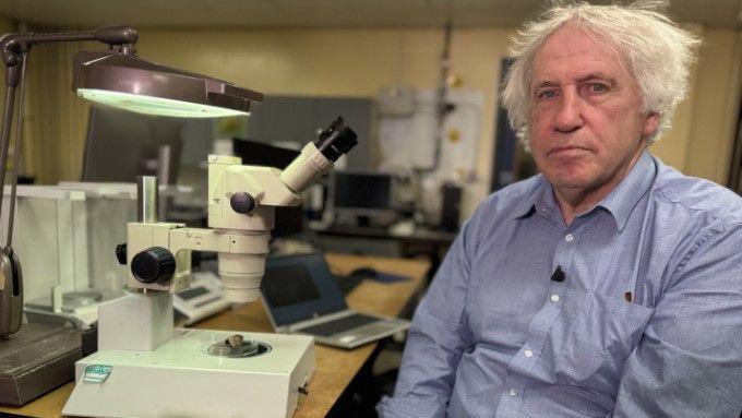 A man with white hair wearing a blue shirt is sat in a lab beside a microscope