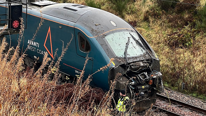 An Avanti West Coast train on a track in a rural setting. The front of the train is smashed and covered in mud