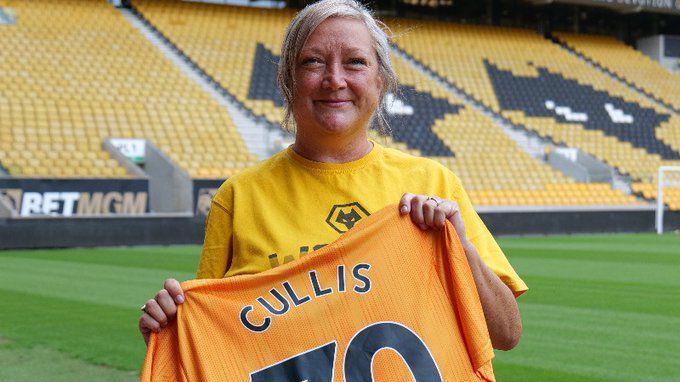 A woman holds an orange football t-shirt with word Cullis and the number 70. She is standing on the pitch at a stadium.