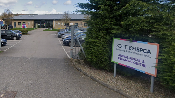 A sign reading "Scottish SPCA, Scotland's Animal Welfare Charity" is mounted on two posts outside the metal gates to a rescue centre in Drumoak. Vehicles including an SSPCA van are parked outside the building.