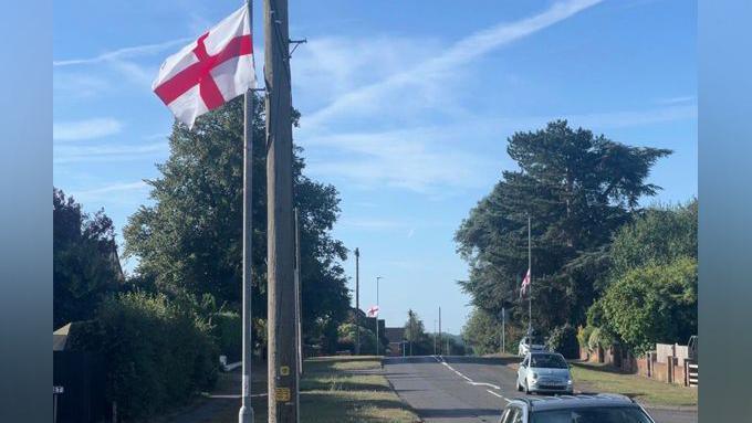 A red and white St George's Flag is tied to a lamppost beside a residential road, and there are two more in the background. There are a couple cars in the frame. There is a blue sky.