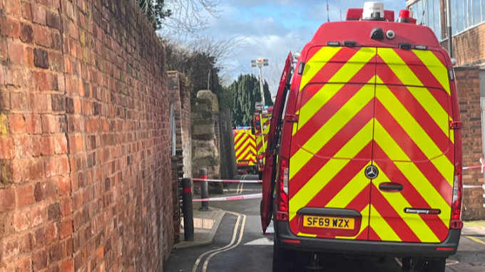 A row of ambulances and fire engines can be seen in an alleyway. There is a long and quite high brick wall either side of the vehicles.