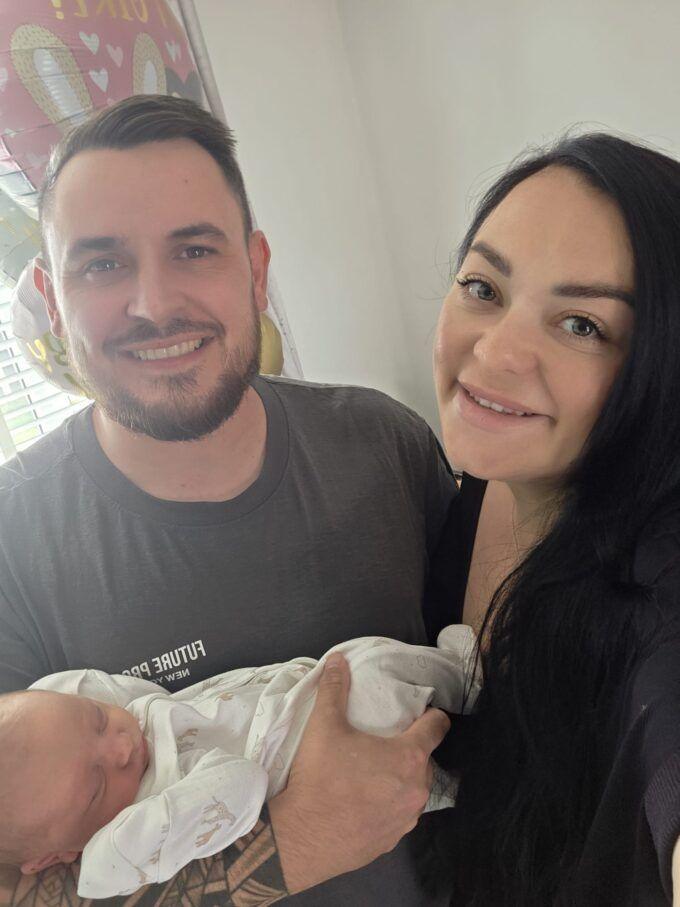 Man with short dark hair and beard with a dark top with a baby wrapped in white and a woman with brown hair and a dark top. They are standing in a living room.