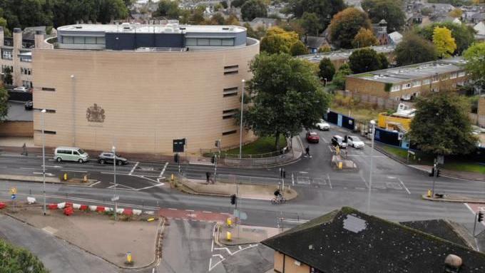 An aerial view of the Cambridge Crown Court building and the roads surrounding it. Cars can been seen approaching traffic lights in front of the building, which is made of light-coloured bricks and has four storeys.