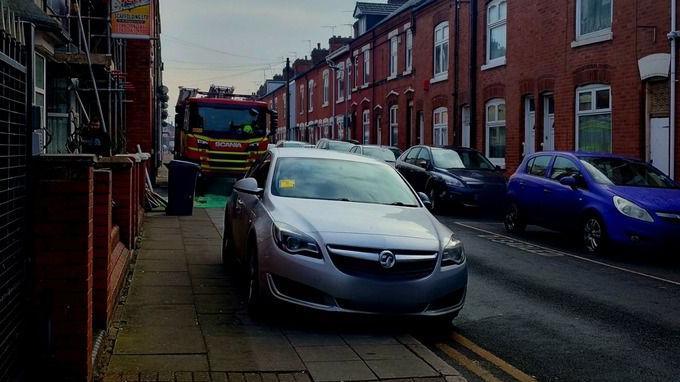 A car on the pavement in a double yellow lined area with a yellow sticker on its windscreen and a fire engine parked nearby