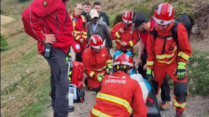 A group of seven emergency workers in red coats with reflective yellow bands, and two in a dark green uniform, along with a person in a baseball cap and grey coat, are stood around a person on a stretcher on a hillside path. Some are kneeling around a person on the stretcher.