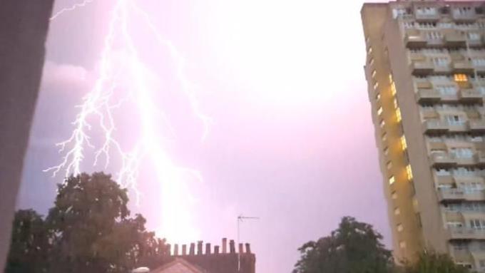 A bright lightning bolt forks across a purple-lit sky above residential buildings, including a tall tower block, during a night-time thunderstorm.