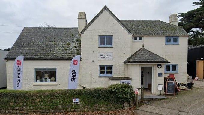 A whitewashed two-storey building has pop up signs saying 'Leach Pottery' and 'shop' and a sign on the property itself saying 'The Leach Pottery'.