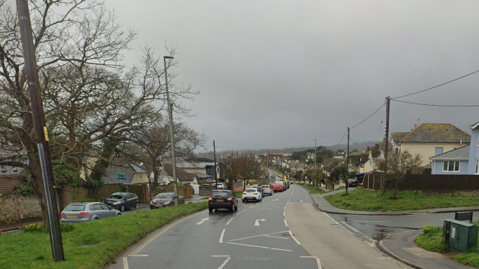 Exeter Road in Dawlish. Parked cars and cars in a traffic queue, grass verges and houses at the side of the road.  Cloudy skies.