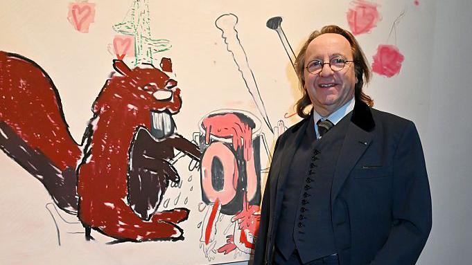 Bill Pallot poses for a photo next to an artwork at a gallery exhibition in Paris in April. He wears a three-piece dark suit, round-rimmed spectacles and has shoulder-length hair.