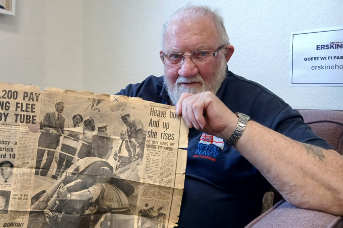 Simon Murdoch holds up a newspaper showing people looking at a giant tortoise. Simon has white short hair and a white beard and wears a dark Royal Navy polo shirt