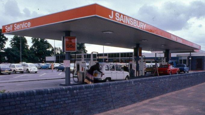 A petrol station behind a low wall in about 1975. It has a canopy roof branded in orange with self service, J Sainsbury and petrol station written in white lettering. Beneath the canopy a woman in a floor-length skirt can be seen putting petrol into a white car. Beyond the station is a carpark and a supermarket, with summer trees in leaf