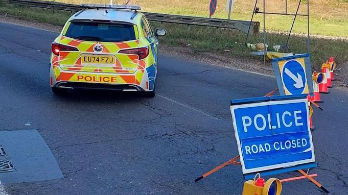 A police car parked on a road with a sign that says "Police Road Closed" and there are traffic cones also on the road 