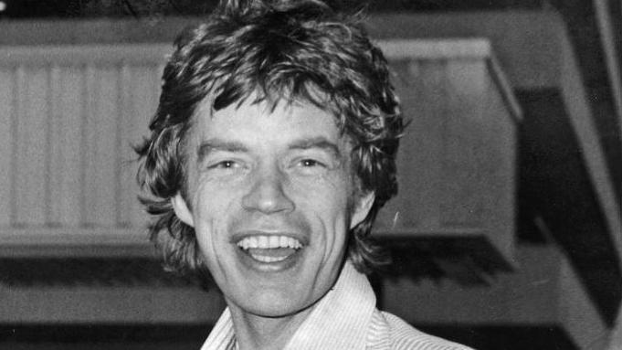 Mick Jagger in a stripe shirt is captured in a black and white photograph at Heathrow Airport