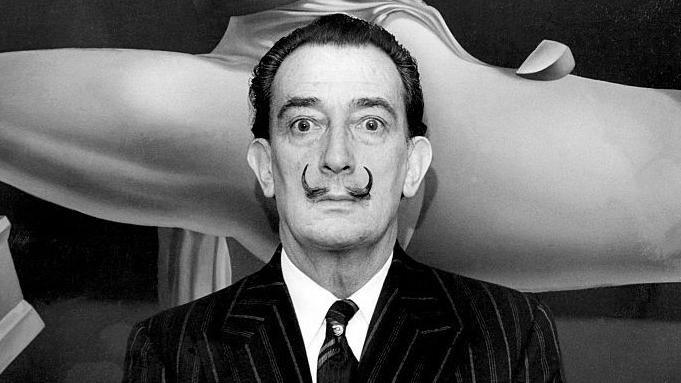 Salvador Dali at the opening of an exhibition of his work in New York. He is wearing a striped jacked and dark tie and has a black moustache which is shaped into two fine points, curling up to his nose. The photograph is black and white.