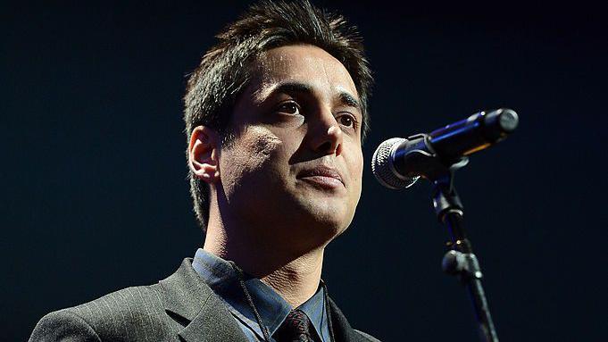 Kianoosh Sanjari speaks into a microphone onstage. He has short dark hair and wears a grey blazer and blue shirt. 