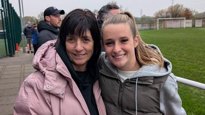 A woman with a dark bob and wearing a pink puffer jacket stands with footballer Ella Toone - a young women with long blond hair in a ponytail. They are standing in front of an empty football pitch.