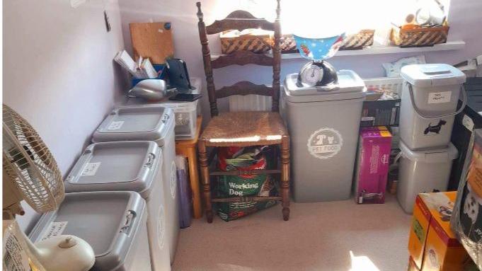 Wendy Isaacs' spare room which is used to store supplies for the pet food bank. Several large grey plastic containers can be seen against the wall. Each one is labelled with its contents. Smaller containers can also be seen, as well as boxes of dog food and cat food. 