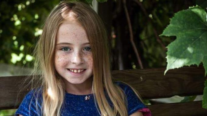 Cimarron Thomas is staring into the camera. She has dark blonde hair and blue eyes. She is wearing a blue dress with short sleeves. She is sitting on a bench with trees in the background