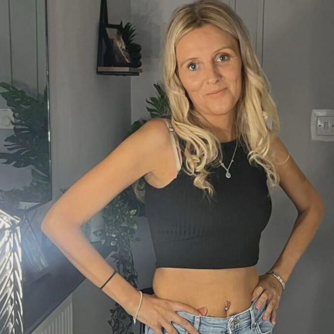 Carol O'Reilly has long, blonde hair and is wearing a black crop top and faded blue jeans. She is standing by a mirror and is smiling at the camera