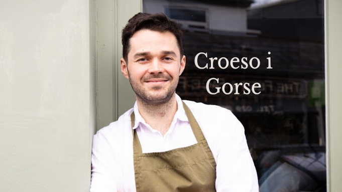 Tom Waters stands outside the door to his restaurant with a sign that says 'Croeso i Gorse' 