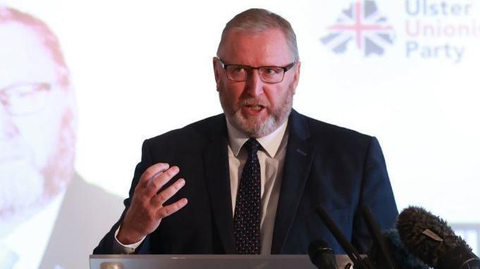 Doug Beattie standing at a podium and speaking at the Ulster Unionist Party manifesto launch. He is wearing a blue suit jacket, tie and white shirt.  