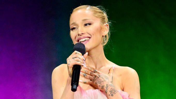 Ariana Grande holding a black microphone smiling and singing