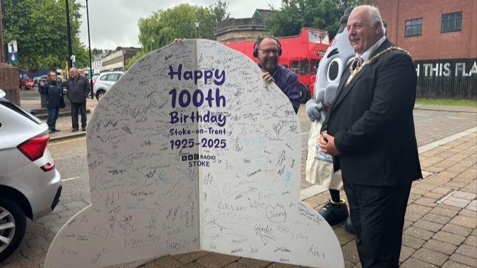 A card with lots of signatures on it and the words "Happy 100th Birthday Stoke-on-Trent 1925-2025 BBC Radio Stoke" - a man's head pokes out from behind the card and a man in a suit stands to the right of it