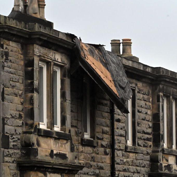 A two-storey sandstone building is shown with part of its roof blown off and hanging over a window on the top floor 