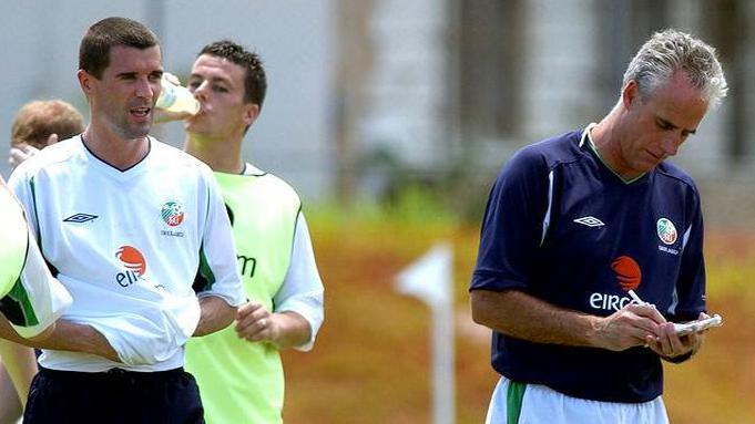 Manager Mick McCarthy (right) and Roy Keane pictured on the pitch at a training session in Saipan. Mick has short white hair and is writing on a small notepad. He is wearing a dark blue Ireland team football shirt and white Ireland team shorts. Roy has short dark hair and is wearing a white Ireland team football shirt and dark Ireland team shorts.