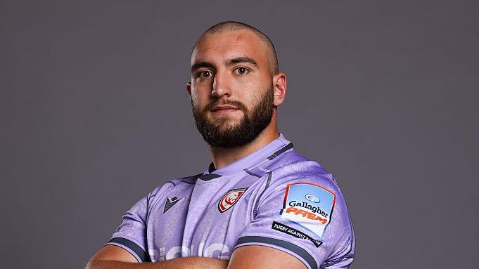 Archie McArthur standing looking at the camera with his arms folded. He is wearing purple Gloucester kit