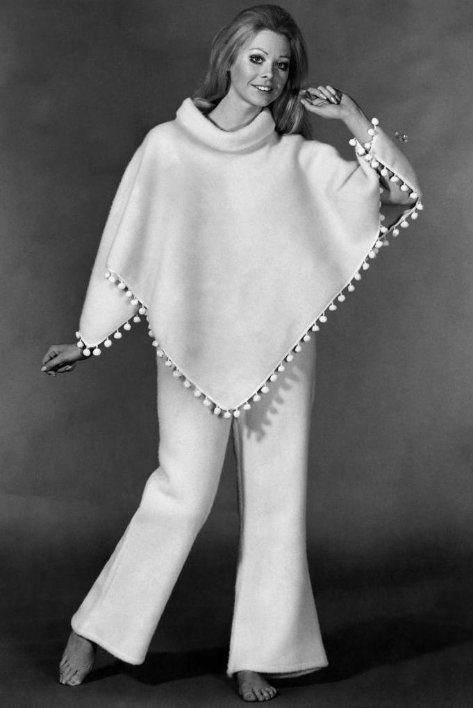 Black and white advertising photo of a woman wearing "poncho pyjamas" - a white poncho trimmed with bobbles and flared trousers. The model has bare feet, heavy eye makeup, and one arm aloft. 