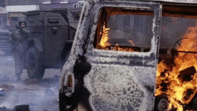 A burning car in the foreground of an historical street scene in Northern Ireland. In the background, a British soldier and Army vehicles