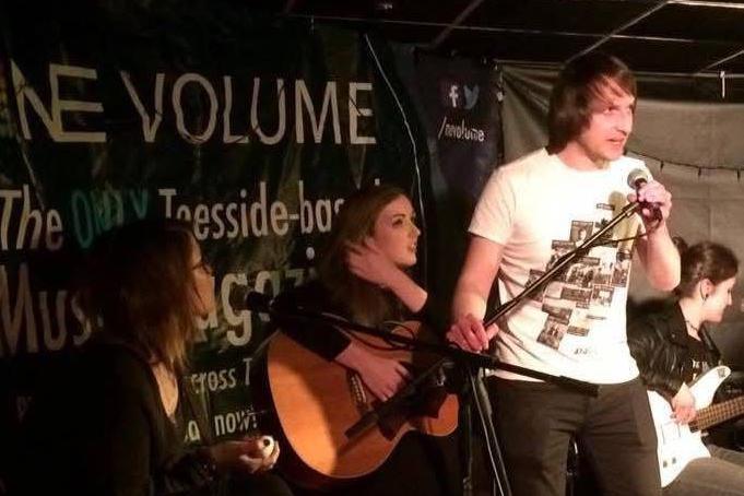 Lee Allcock stands on stage and is speaking to a microphone. There are two musicians sitting behind hime. A banner reading: "NE Volume" is erected behind them all.