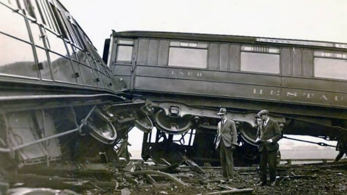 A black and white image of a train crash from 1928 with two carriages off the line. Two men wearing bowler hats stand in the wreckage