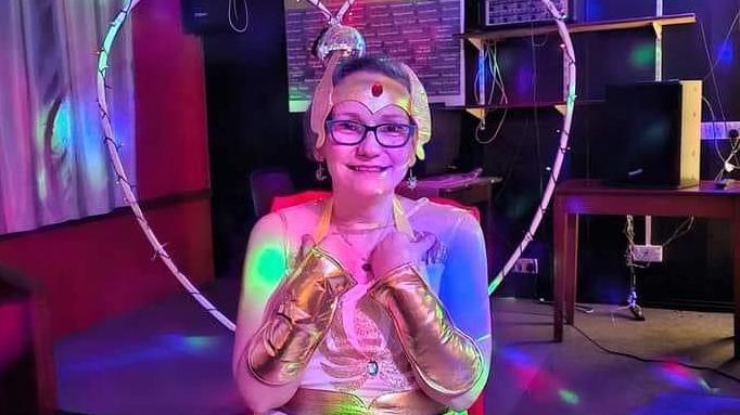 Jenni is in fancy dress, wearing a gold headdress, with a red stone in the middle. She has gold sleeves and blue spectacles. She is standing in front of a heart-shaped structure decorated with fairy lights. 